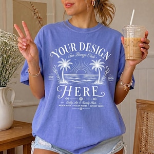 May include: A periwinkle blue short-sleeve t-shirt featuring white graphic text that reads YOUR DESIGN HERE, Sea Breeze Club, EST. 2026, and Salty Air & Sandy Hair. The design includes a sun rising over the ocean between two palm trees.