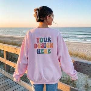 以下が含まれることがあります： レトロなフォントで「YOUR DESIGN HERE」と書かれた、明るいピンク色のスウェットシャツ。画像はスウェットシャツの背面を示しており、背景にはビーチと海があります。モデルはヘアクリップを着用しています。