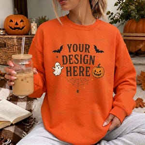 May include: An orange crewneck sweatshirt featuring a black graphic print that reads YOUR DESIGN HERE, accompanied by illustrations of a ghost, a jack-o'-lantern, two bats, and a spider web. The sweatshirt has long sleeves and a ribbed neckline.