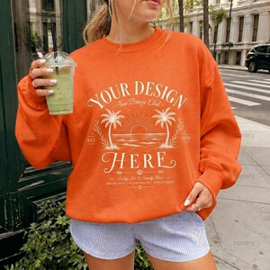 May include: An orange long-sleeve sweatshirt featuring a white graphic print of a sunset, palm trees, and the text "YOUR DESIGN HERE Sea Breeze Club EST. 2026" with additional smaller text including "Beach Days" and "Sandy Hair".