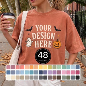 Puede incluir: Una camiseta de manga corta color óxido con el texto YOUR DESIGN HERE en blanco, rodeado de siluetas de murciélagos negros, un fantasma blanco y una calabaza naranja. Un distintivo negro muestra el texto 48 COLORS sobre una paleta de colores.