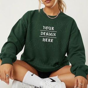 Puede incluir: Una sudadera verde oscuro con el texto "YOUR DESIGN HERE" en blanco. La sudadera tiene mangas largas y cuello redondo. La modelo lleva calcetines y zapatillas blancas.
