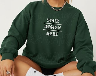 May include: A dark green sweatshirt with the text "YOUR DESIGN HERE" in white. The sweatshirt has long sleeves and a crew neck. The model is wearing white socks and sneakers.