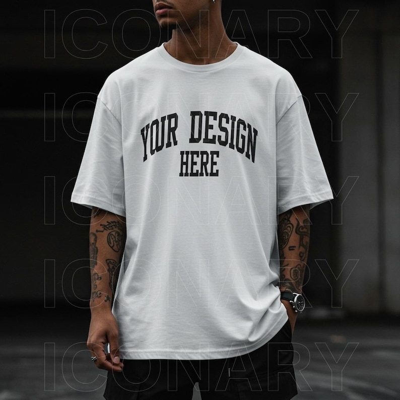 A fashion model, styled in contemporary streetwear, stands against a dark, urban backdrop. He wears a plain white oversized t-shirt with the word "YOUR DESIGN HERE" printed in a bold, black, collegiate-style font.   The t-shirt is a blank canvas design mock-up; the text is a light gray. The man's skin tone is medium, and he has short, dark hair and visible tattoos on his arms. He is wearing dark-colored, loose-fitting trousers, and has a watch on his wrist.