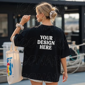 Könnte beinhalten: Schwarzes übergroßes T-Shirt mit dem Text "YOUR DESIGN HERE" in Weiß. Die Person hält eine Tragetasche aus Segeltuch mit einem farbenfrohen abstrakten Design. Die Person trägt ein Scrunchie und goldene Creolen.
