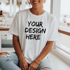 Könnte beinhalten: Weißes T-Shirt mit dem schwarzen Text "YOUR DESIGN HERE". Das T-Shirt hat kurze Ärmel und einen Rundhalsausschnitt. Die Person trägt eine blaue Jeans und ein goldenes Armband. Das Bild ist bei natürlichem Licht aufgenommen.