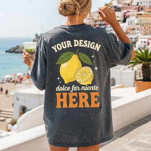 以下が含まれることがあります： レモン柄と「YOUR DESIGN dolce far niente HERE」の文字が入ったダークグレーのTシャツ。デザインは丸ごとのレモンと、緑の葉が付いたレモンスライスが特徴です。ゆったりとしたフィット感のシャツです。