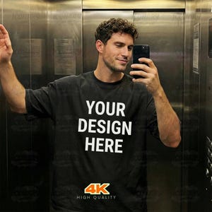 Puede incluir: Camiseta negra con el texto "YOUR DESIGN HERE" en letras blancas. La camiseta también presenta un logotipo "4K HIGH QUALITY" en la parte inferior. La persona de la imagen se está haciendo un selfie.