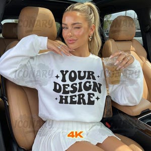 May include: A white sweatshirt with the text "YOUR DESIGN HERE" in black, paired with a white pleated skirt. The image is labeled "4K HIGH QUALITY". The person is holding a coffee cup.