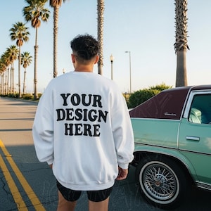 Peut inclure: Sweat-shirt blanc avec le texte "YOUR DESIGN HERE" en lettres noires au dos. La personne se tient sur une route à côté d'une voiture verte vintage. Des palmiers bordent la rue sous un ciel clair.