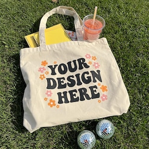 Puede incluir: Una bolsa de lona beige con texto negro que dice "YOUR DESIGN HERE" rodeada de flores naranjas y rosas. La bolsa est&aacute; sentada sobre hierba verde con dos bolas de discoteca plateadas.