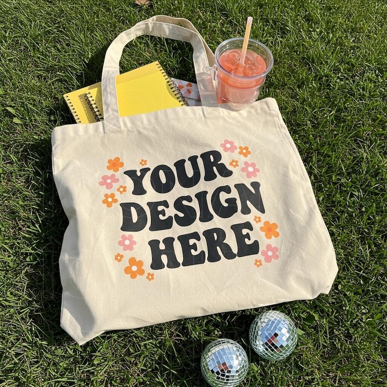 Puede incluir: Una bolsa de lona beige con texto negro que dice "YOUR DESIGN HERE" rodeada de flores naranjas y rosas. La bolsa est&aacute; sentada sobre hierba verde con dos bolas de discoteca plateadas.