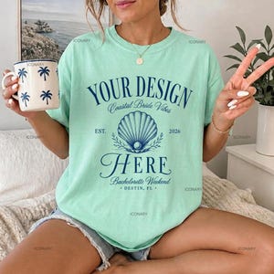 May include: A light green t-shirt featuring dark blue text that reads YOUR DESIGN, Coastal Bride Vibes, EST. 2026, HERE, Bachelorette Weekend, DESTIN, FL, and a central seashell graphic. A white mug with blue palm tree prints is held in one hand.