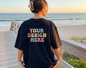 Puede incluir: Camiseta negra con el texto "YOUR DESIGN HERE" en una fuente retro colorida. La camiseta la lleva una persona en un paseo marítimo de madera con vistas al mar al atardecer. La camiseta tiene un corte holgado.