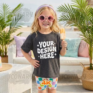 Puede incluir: Una niña con una camiseta gris oscuro con el texto "YOUR DESIGN HERE" en blanco. También lleva pantalones cortos a cuadros coloridos, gafas de sol rosas con forma de flor y una diadema morada. Sostiene un helado de naranja.