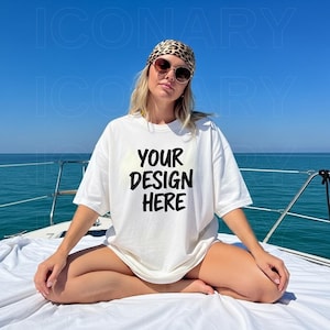 May include: A white oversized t-shirt with the text "YOUR DESIGN HERE" in black. The person is wearing sunglasses and a leopard print headscarf, sitting on a boat with the ocean and blue sky in the background.