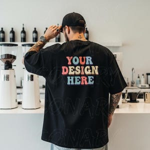 May include: Black oversized t-shirt with the text "YOUR DESIGN HERE" in a colourful retro font. The shirt is worn by a person in a black cap and watch. The background includes coffee machines and bottles.