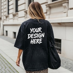 以下が含まれることがあります： 白で「YOUR DESIGN HERE」の文字が入った黒のオーバーサイズTシャツ。モデルはレオパード柄のスカートを履き、黒のトートバッグを持っています。シャツは半袖で、クルーネックです。