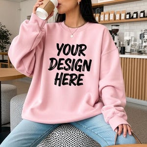 Op de afbeelding: Een lichtroze sweatshirt met de tekst "YOUR DESIGN HERE" in het zwart. De sweatshirt heeft een ronde hals. De persoon op de foto draagt een blauwe spijkerbroek en houdt een koffiekopje vast.