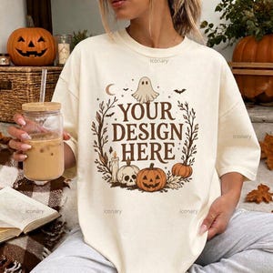 Puede incluir: Una camiseta color crema con un estampado gráfico marrón de un fantasma, una calavera, una calabaza, una vela y una luna creciente rodeados por una corona de hojas. El texto "YOUR DESIGN HERE" está impreso en el centro en fuente negrita.