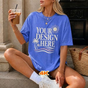 May include: A blue t-shirt with the text "YOUR DESIGN HERE" in white, featuring a sun, palm tree, and wave design. The shirt is worn by a person holding a beverage in a clear cup. The image also shows a straw bag and white trainers.