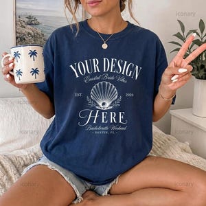 May include: A navy blue t-shirt featuring white text that reads YOUR DESIGN HERE, Coastal Bride Vibes, EST. 2026, Bachelorette Weekend, DESTIN, FL. A white seashell graphic is centered on the chest. A white mug with blue palm tree prints is held.