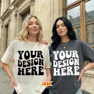 May include: Two women wearing t-shirts with the text "YOUR DESIGN HERE" in a retro font. One shirt is cream-colored, the other is dark gray. Both shirts have short sleeves and a relaxed fit. The image is taken outdoors.
