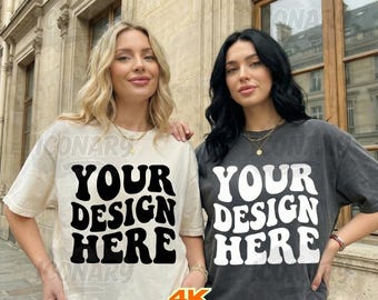 May include: Two women wearing t-shirts with the text "YOUR DESIGN HERE" in a retro font. One shirt is cream-colored, the other is dark gray. Both shirts have short sleeves and a relaxed fit. The image is taken outdoors.