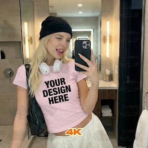 May include: A light pink crop top with the text "YOUR DESIGN HERE" in black. The person in the image is wearing a black beanie and white headphones. The person is holding a black phone.
