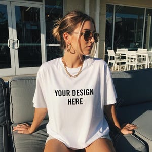 Könnte beinhalten: Weißes T-Shirt mit dem Text "YOUR DESIGN HERE" in Schwarz. Das Model trägt goldfarbenen Schmuck, darunter eine Halskette und Creolen. Das Model trägt auch eine Sonnenbrille.