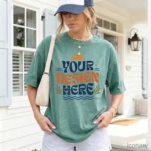 May include: A green t-shirt with the text "YOUR DESIGN HERE" in orange and blue. The shirt has a retro design with a sun, waves, and leaves. The person is wearing a blue and white baseball cap and a white handbag.