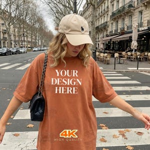 Puede incluir: Una camiseta naranja quemado con el texto "YOUR DESIGN HERE" en letras blancas. La camiseta se combina con una gorra de béisbol beige con un logotipo negro. Un bolso negro con una correa de cadena cuelga del hombro.