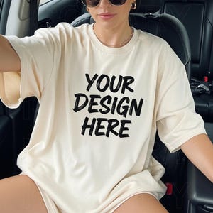 Może przedstawiać: Kremowa koszulka oversize z napisem "YOUR DESIGN HERE" pogrubioną, czarną czcionką. Koszulka ma krótkie rękawy i luźny krój. Zdjęcie przedstawia noszoną koszulkę.