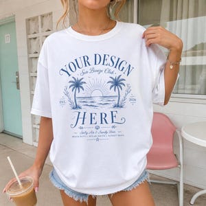 以下が含まれることがあります： YOUR DESIGN HERE、Sea Breeze Club、EST. 2026、Salty Air & Sandy Hairというテキストが青色でプリントされた白い半袖Tシャツ。デザインには2本のヤシの木、太陽、海の波のイラストが描かれています。