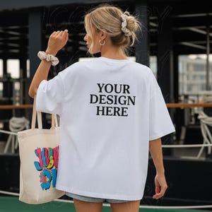 以下が含まれることがあります： 背中に黒で「YOUR DESIGN HERE」とプリントされた白いオーバーサイズのTシャツ。カラフルなグラフィックのベージュのトートバッグが左手に持たれています。シュシュとフープイヤリングを着用しています。