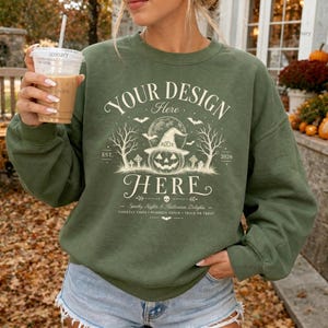 Puede incluir: Una sudadera de cuello redondo color verde oliva con un gráfico en color crema de una calabaza con sombrero de bruja, rodeada de árboles desnudos, murciélagos y lápidas. El texto dice YOUR DESIGN HERE, EST. 2026, Spooky Nights & Halloween Delights.