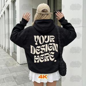 以下が含まれることがあります： クリーム色のレトロなフォントで「YOUR DESIGN HERE」という文字が入った黒いパーカー。ベージュの野球帽と白いスカートを着用。右側には黒いショルダーバッグがかかっています。