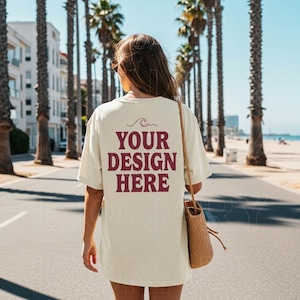 May include: A woman wearing a white t-shirt with the text "YOUR DESIGN HERE" in maroon. She is walking down a street lined with palm trees and a beach in the background. She is carrying a woven straw bag.