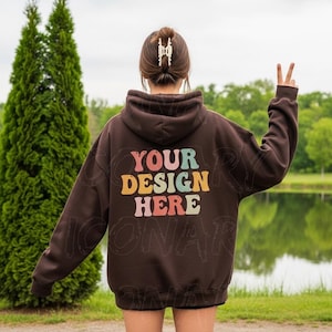 Puede incluir: Una sudadera con capucha marrón con el texto "YOUR DESIGN HERE" en letras retro coloridas. La sudadera con capucha tiene una capucha y mangas largas. La persona está de pie afuera cerca de un lago.