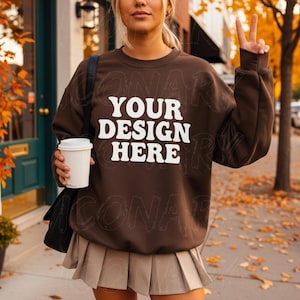 Op de afbeelding: Een bruine sweatshirt met de tekst "YOUR DESIGN HERE" in het wit. De persoon draagt een beige geplooide rok en houdt een witte koffiebeker vast. De achtergrond omvat een deur, herfstbladeren en een stoep.