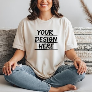 May include: A cream-coloured t-shirt with the text "YOUR DESIGN HERE" in black, centred on the front. The short-sleeved shirt is made of a soft material. The model is wearing blue jeans.