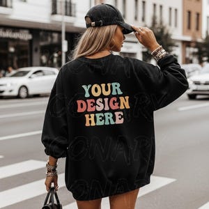 以下が含まれることがあります： カラフルなレトロフォントで「YOUR DESIGN HERE」と書かれた黒いスウェットシャツ。黒い野球帽をかぶり、黒いハンドバッグを持っています。背景には、車や建物のある街並みが写っています。