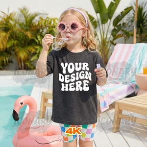 Puede incluir: Un niño lleva una camiseta gris oscuro con el texto "YOUR DESIGN HERE" en blanco, soplando burbujas. Lleva gafas de sol rosas con forma de flor y pantalones cortos coloridos. Un flotador de piscina de flamenco rosa está en el fondo.