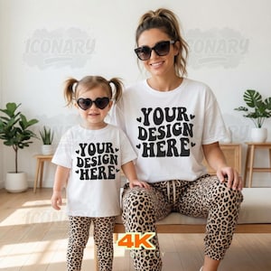 May include: A woman and child in matching white t-shirts printed with "YOUR DESIGN HERE" in black. The woman wears leopard print trousers and sunglasses. The child wears leopard print leggings and heart-shaped sunglasses. The image is taken indoors.