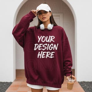 May include: A maroon sweatshirt with the text "YOUR DESIGN HERE" in white. The person in the image is wearing a white baseball cap and white headphones. They are holding a clear plastic cup with a straw.