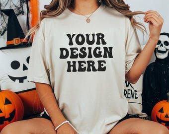 May include: A cream-colored t-shirt with the text "YOUR DESIGN HERE" in black, displayed on a person. The shirt is part of a Halloween-themed setup, with pumpkins, a skull, and decorative pillows in the background.