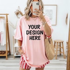 May include: A light pink oversized t-shirt with the text "YOUR DESIGN HERE" in black. The person is wearing red and white striped shorts and carrying a woven tote bag. The background includes a wooden clothing rack and a small wooden stool.