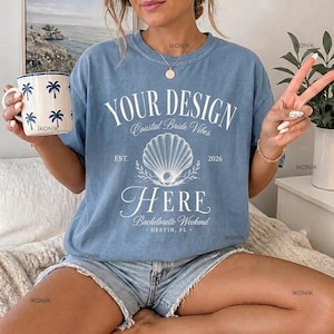Op de afbeelding: Een blauw t-shirt met ronde hals en witte tekst YOUR DESIGN HERE, Coastal Bride Vibes, EST. 2026 en DESTIN, FL, met een centrale afbeelding van een schelp. Het shirt wordt gedragen met een lichtblauwe spijkerbroek en een witte mok met palmbomen.