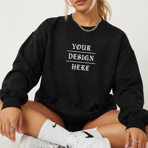 Op de afbeelding: Zwart sweatshirt met de tekst "YOUR DESIGN HERE" in het wit. Het sweatshirt heeft een ronde hals en lange mouwen. Het model draagt witte sokken en sneakers.