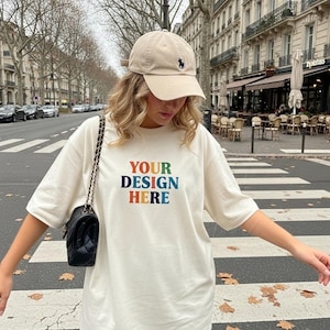 Puede incluir: Camiseta color crema con el texto "YOUR DESIGN HERE" en colores arcoíris. Se lleva una gorra de béisbol beige con un logotipo negro. También se ve un bolso negro con una correa de cadena.